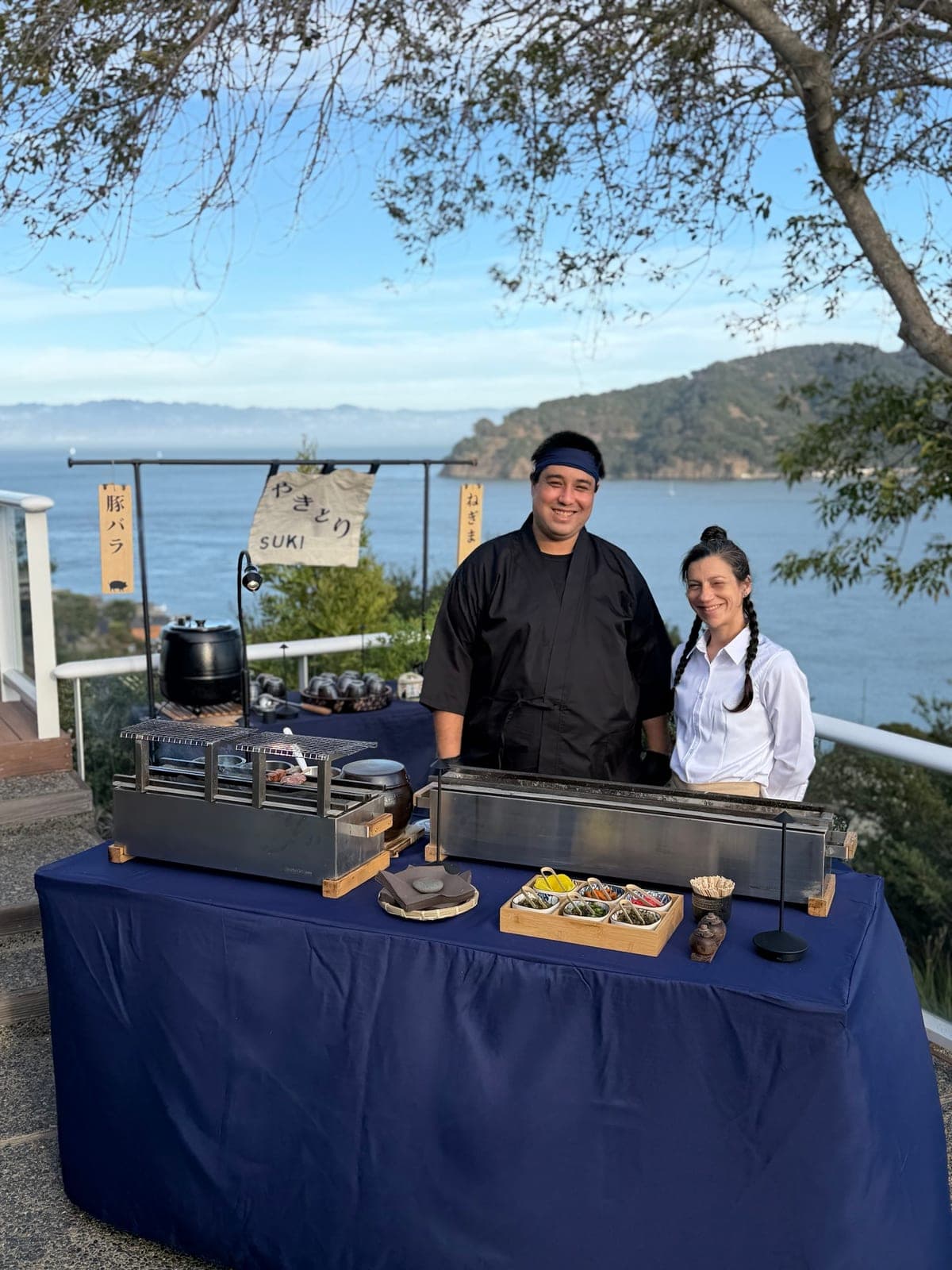 Our chef and a teammate behind two binchotan grills on a navy-covered table, set up on a deck above the bay