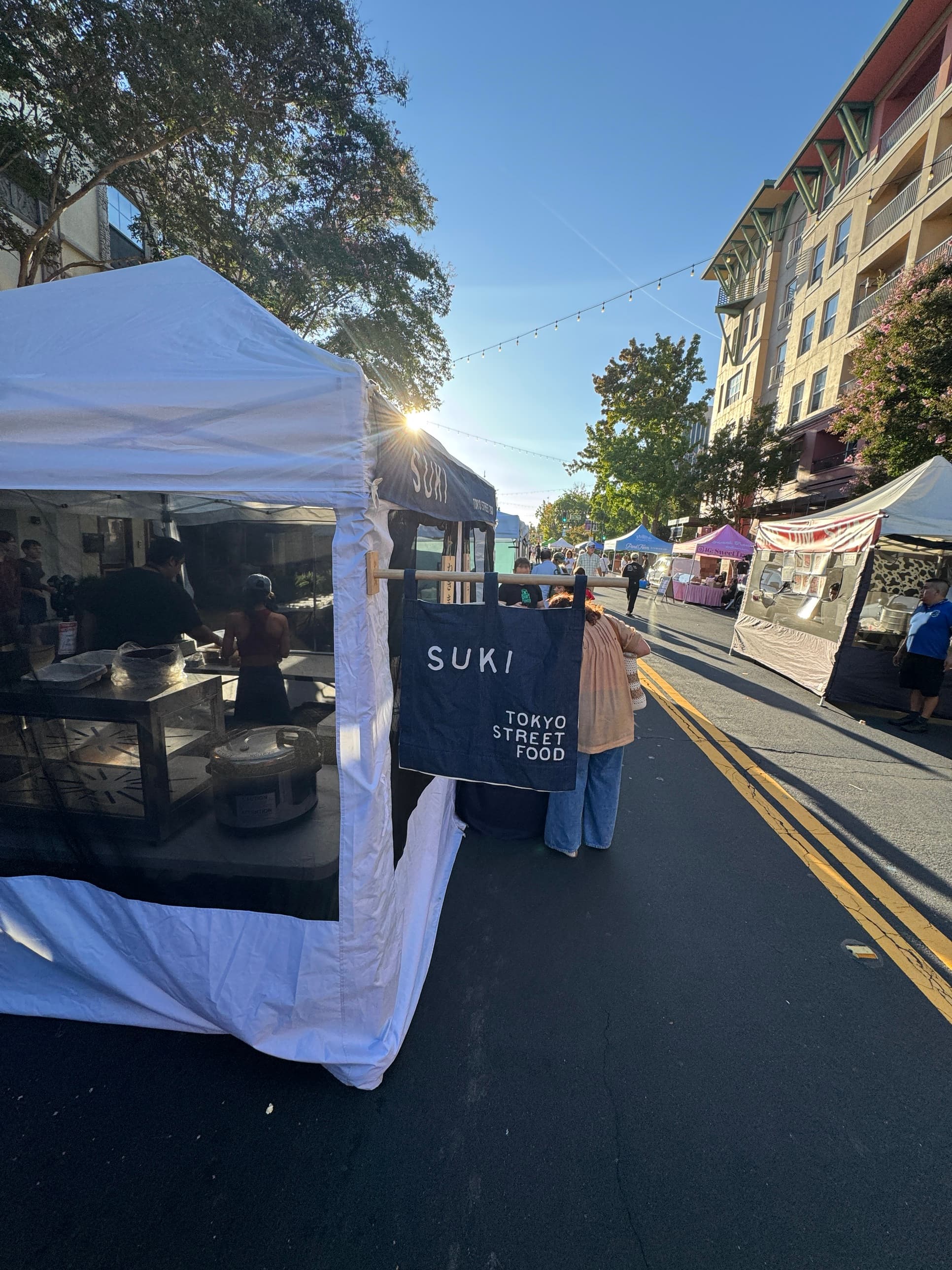 The SUKI tent and navy noren at a downtown street market, string lights overhead