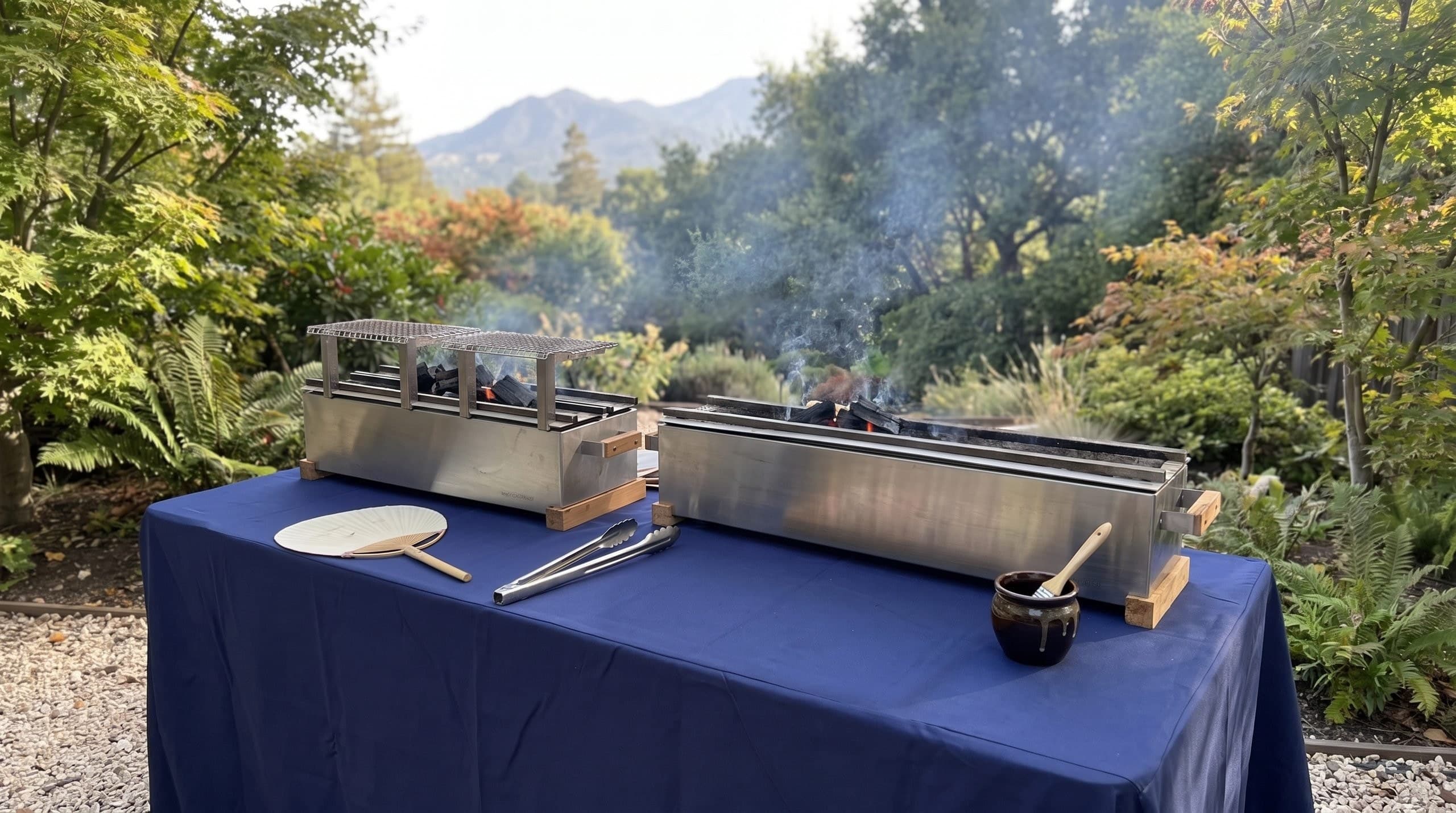 The grill table at the end of the garden path: two stainless binchotan grills on a navy cloth, an uchiwa, tongs, and a pot of tare with a brush